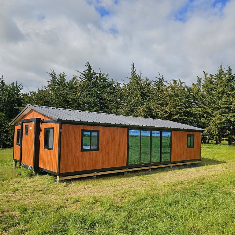 Contemporary wooden tiny home in New Zealand countryside with large glass windows surrounded by lush greenery and blue sky