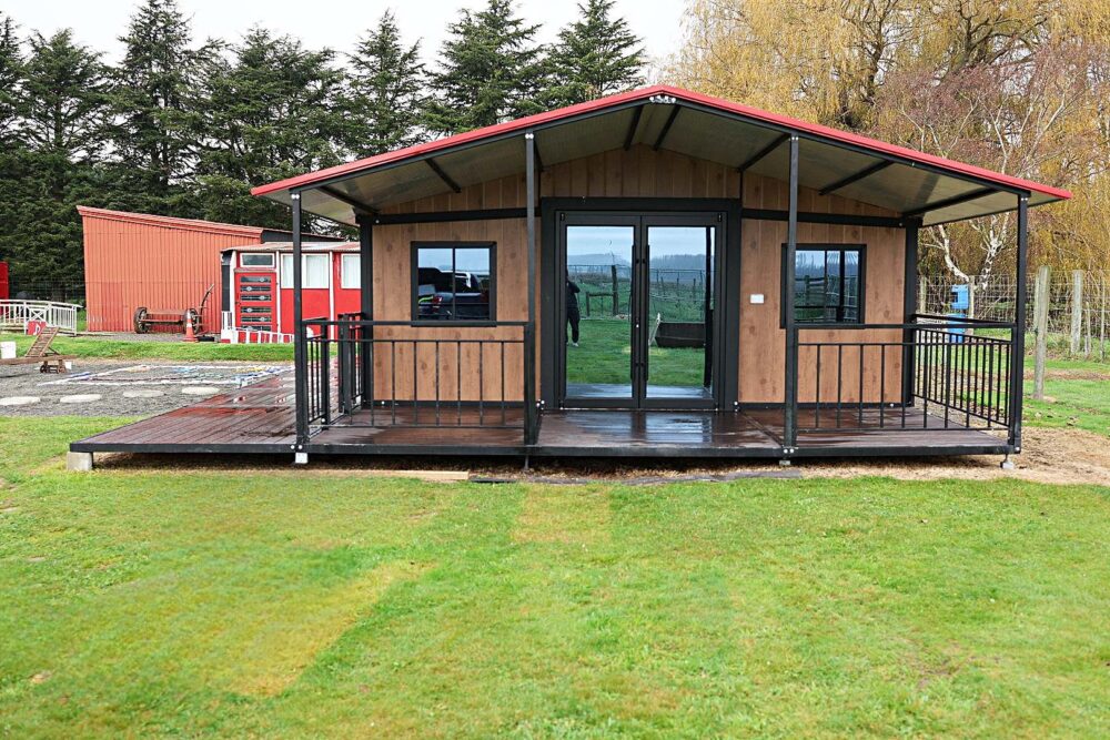 Modern modular home in New Zealand with a gable roof, timber-look cladding, sliding glass doors, and a front deck designed by Portable Dwellings.